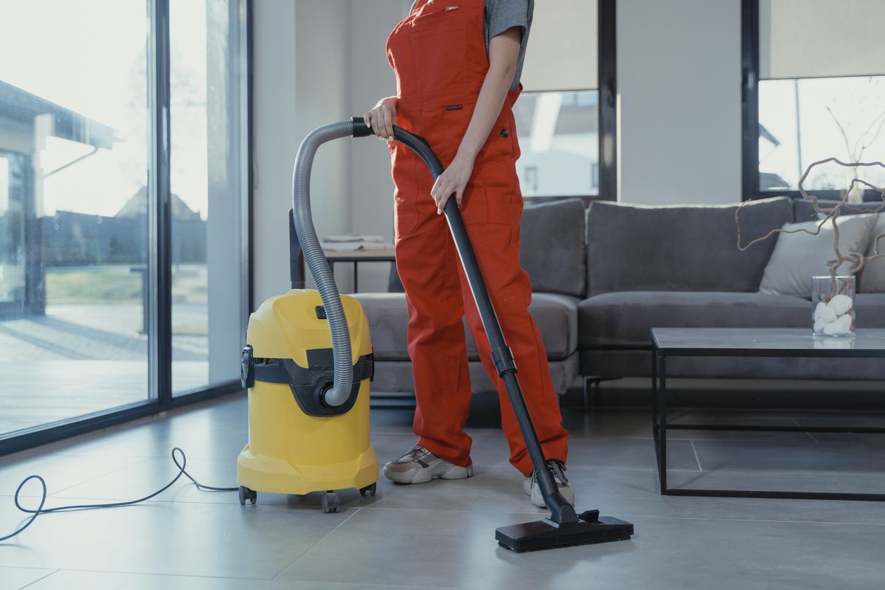 services-01 A professional cleaner in red coveralls vacuuming a contemporary living room with a yellow vacuum cleaner.